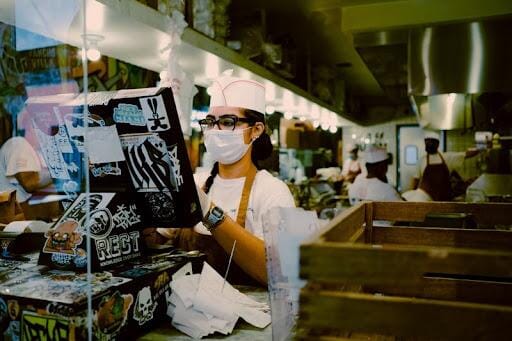 A food service cashier wearing a mask and working behind a clear antimicrobial barrier.
