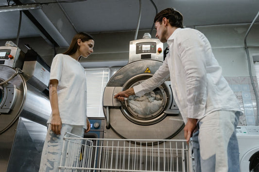 Two people unloading linen from an industrial washing machine into a laundry cart